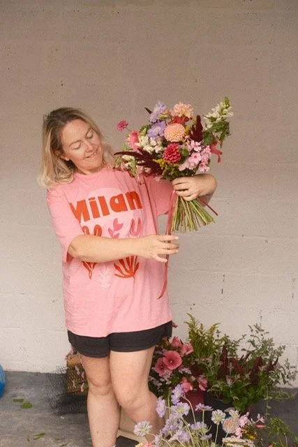 A woman with blonde hair wearing a pink T-shirt and black shorts holding a large colorful bouquet of flowers, standing in front of a concrete wall with potted flowers on the ground.