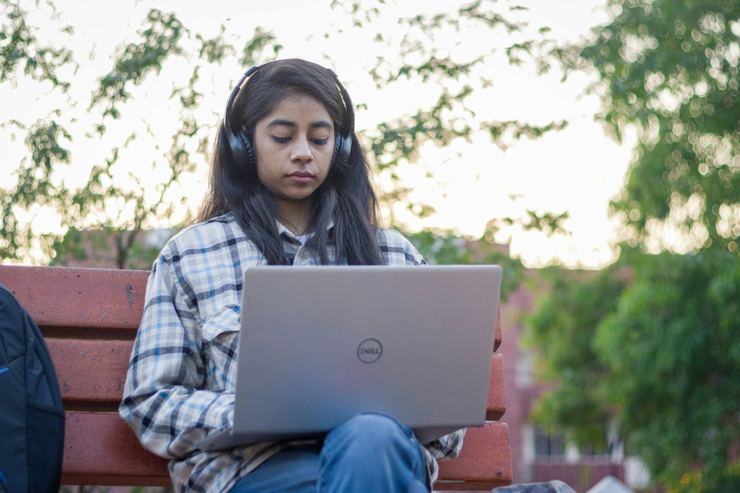 A young woman with long dark hair wearing headphones, a plaid shirt, and jeans, sitting on a red park bench and working on a silver Dell laptop. A navy backpack is next to her, and green trees are in the background.