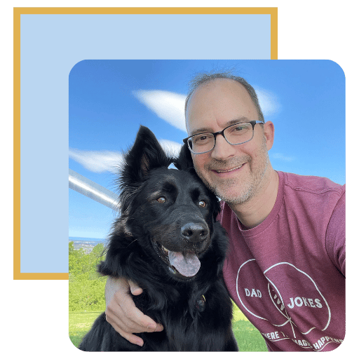 Michael is wearing glasses and a red shirt. He is outdoors with an arm around his black dog.