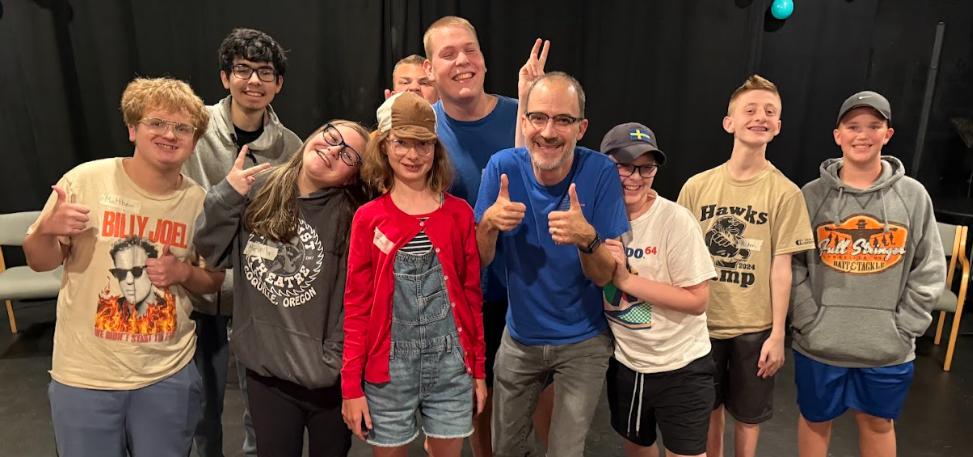 A group of nine people, mostly kids, smiling and posing together in a black room with a black curtain background. They are casually dressed, some making peace signs or thumbs up.