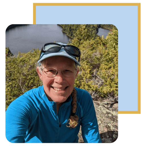 Kelly is wearing glasses, a blue shirt and baseball cap. She is outside with trees and a lake in the background.