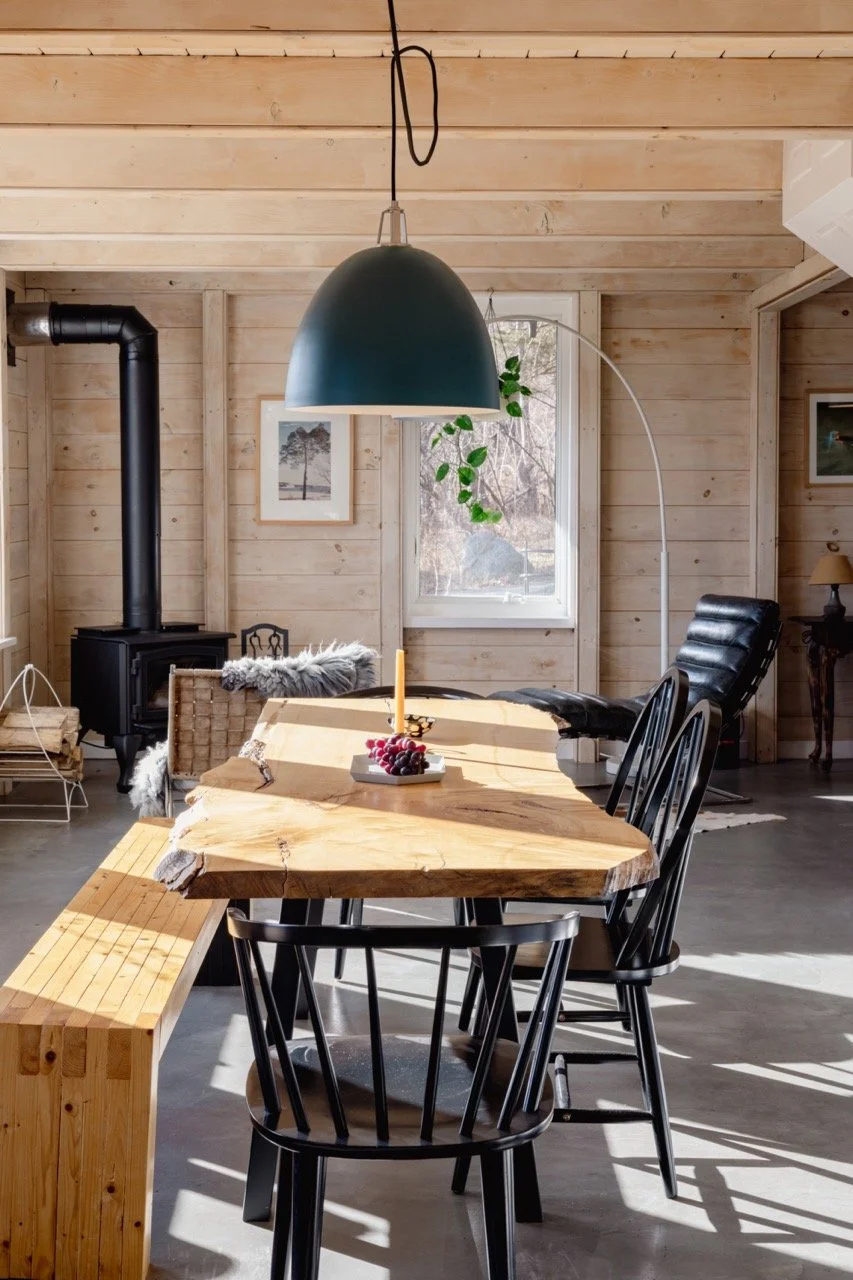 Interior of a cozy wooden cabin dining area with a large natural wood table, black chairs, a modern black pendant light, a black wood stove, a window with natural light, and minimal decor.