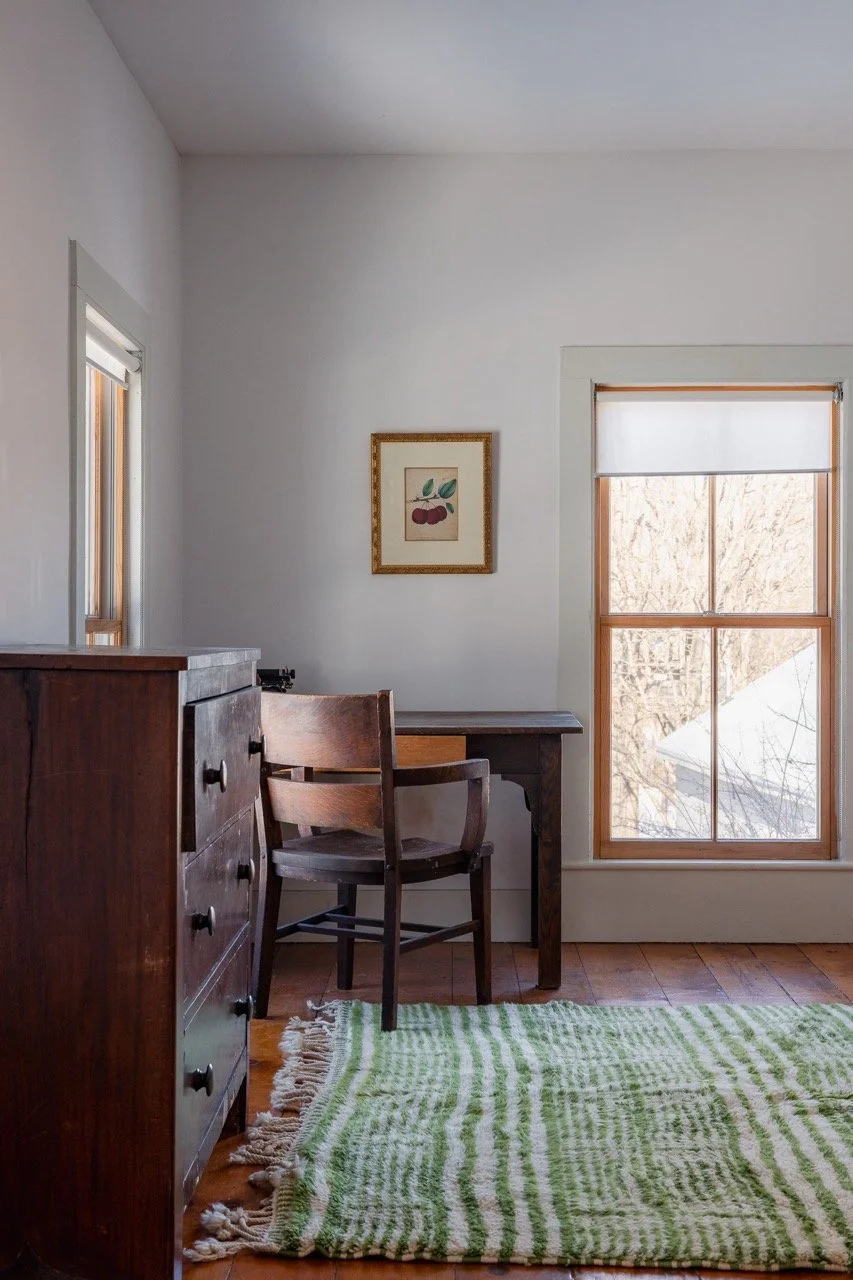 A cozy room corner with a wooden dresser, a small desk, a wooden chair, a window with a view of a snowy outdoor scene, and a green and white patterned rug.