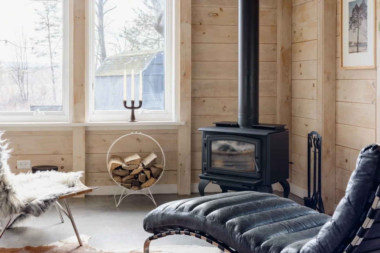 Corner of a cozy living room with a wood stove, a window with a candelabra, a firewood holder filled with logs, a sheepskin-covered chair, and a leather lounge chair.