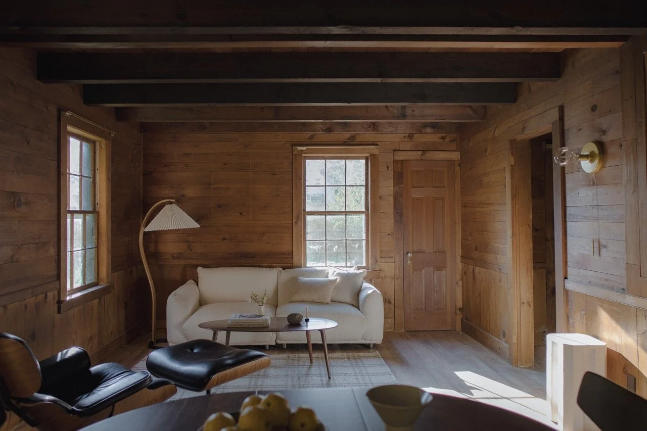 Cozy living room with wooden walls and ceiling beams, featuring a white sofa with pillows, a wooden coffee table, a modern curved floor lamp, and two windows allowing natural light. There is a door and some modern decor elements.