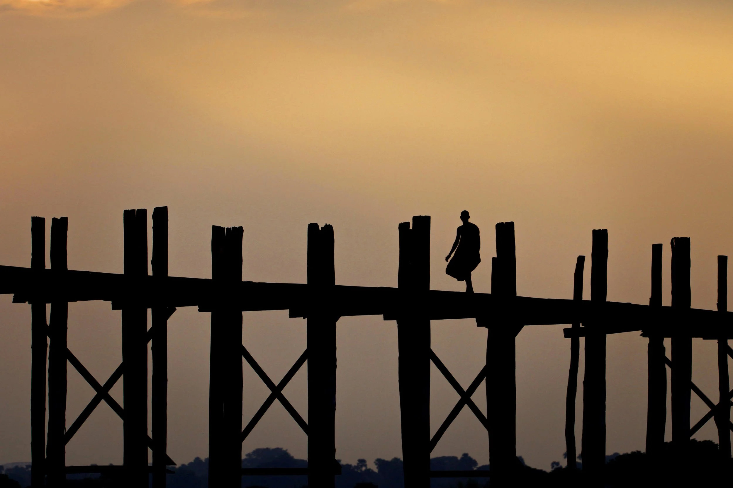 Silhouette of a person walking on a wooden pier at sunset or sunrise, with a colorful sky and calm water below.