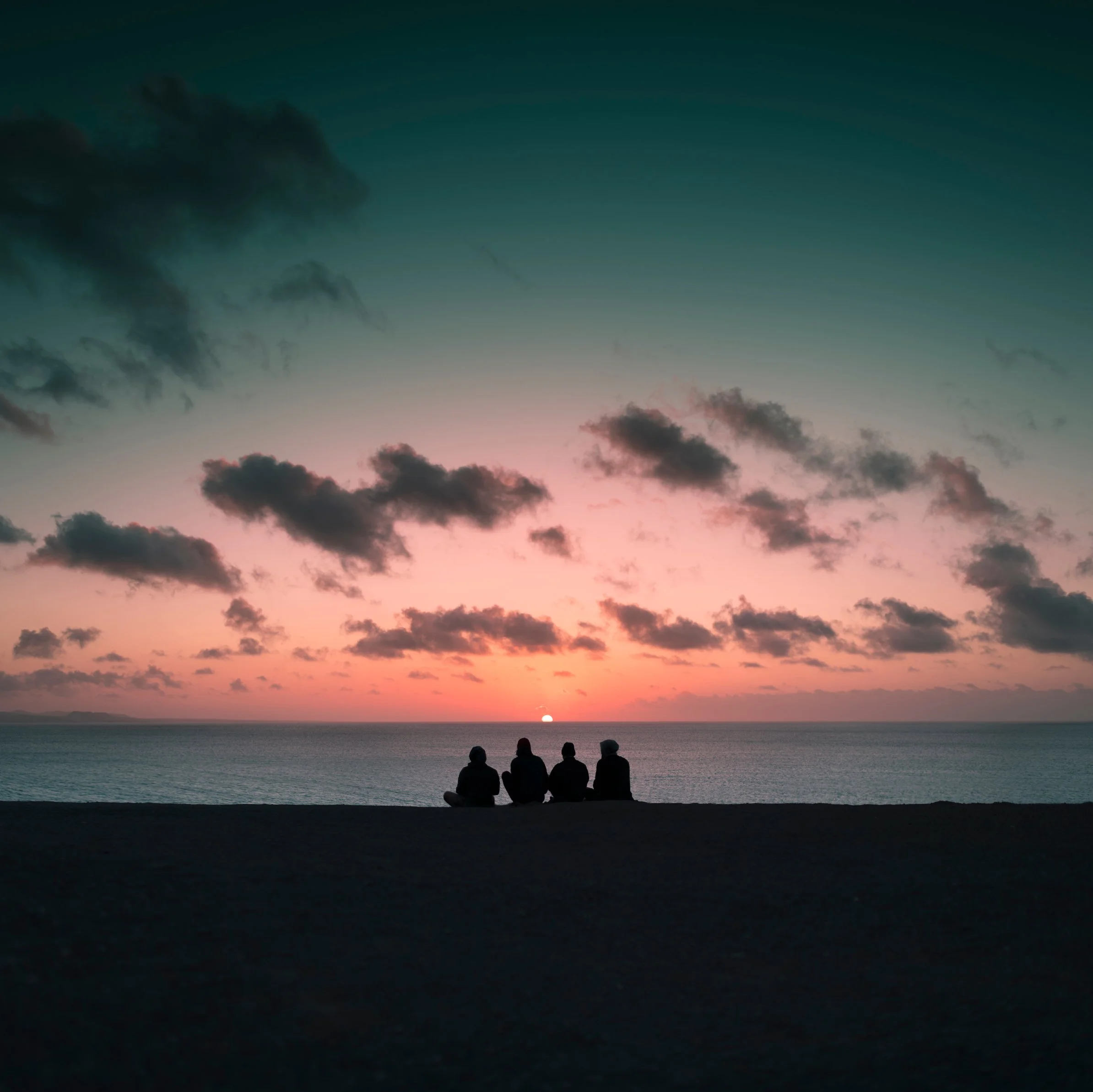 Four people sitting on a beach watching a sunset over the ocean with colorful sky and clouds.