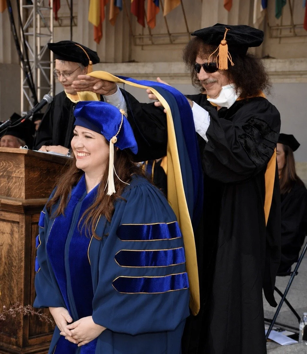 A graduation ceremony where a woman, Dr. Dyan Connelly, in a blue cap and gown is receiving a hood from a man in academic regalia, with other faculty members visible in the background.
