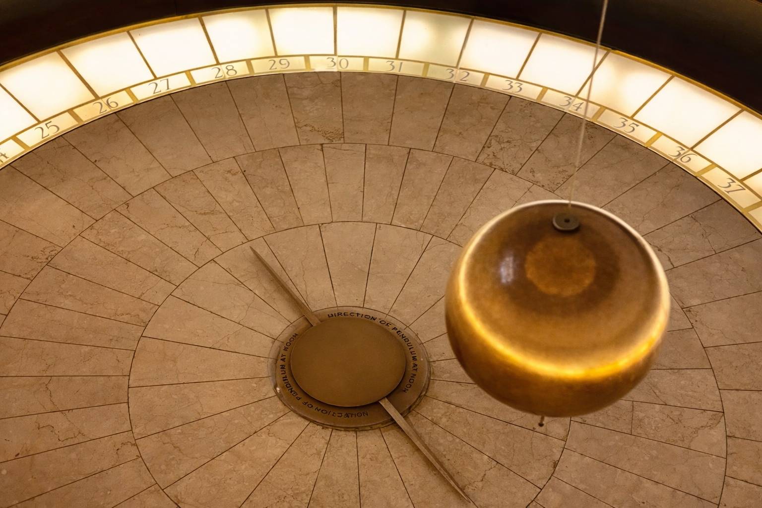 Close up of Griffith Observatory Pendulum, a large, circular clock face with an ornate golden pendulum swinging in the center. The clock face has a marble-like surface with division lines and numbered segments from 25 to 37 at the top.