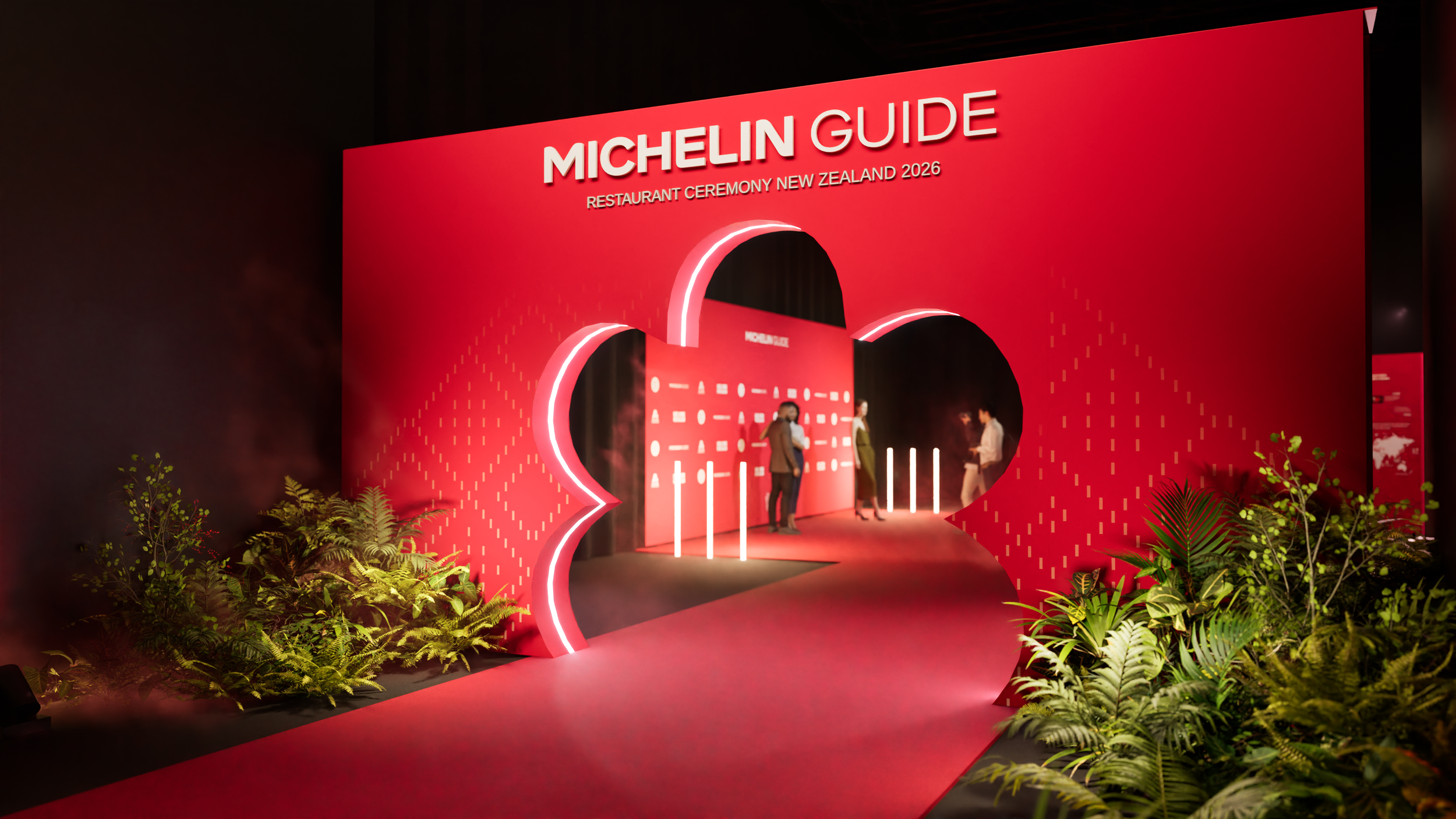 Red stage decorated with plants in front, and a large red backdrop with the text 'Michelin Guide Restaurant Ceremony New Zealand 2026.' There are three people on stage, and bright lights illuminating the scene.