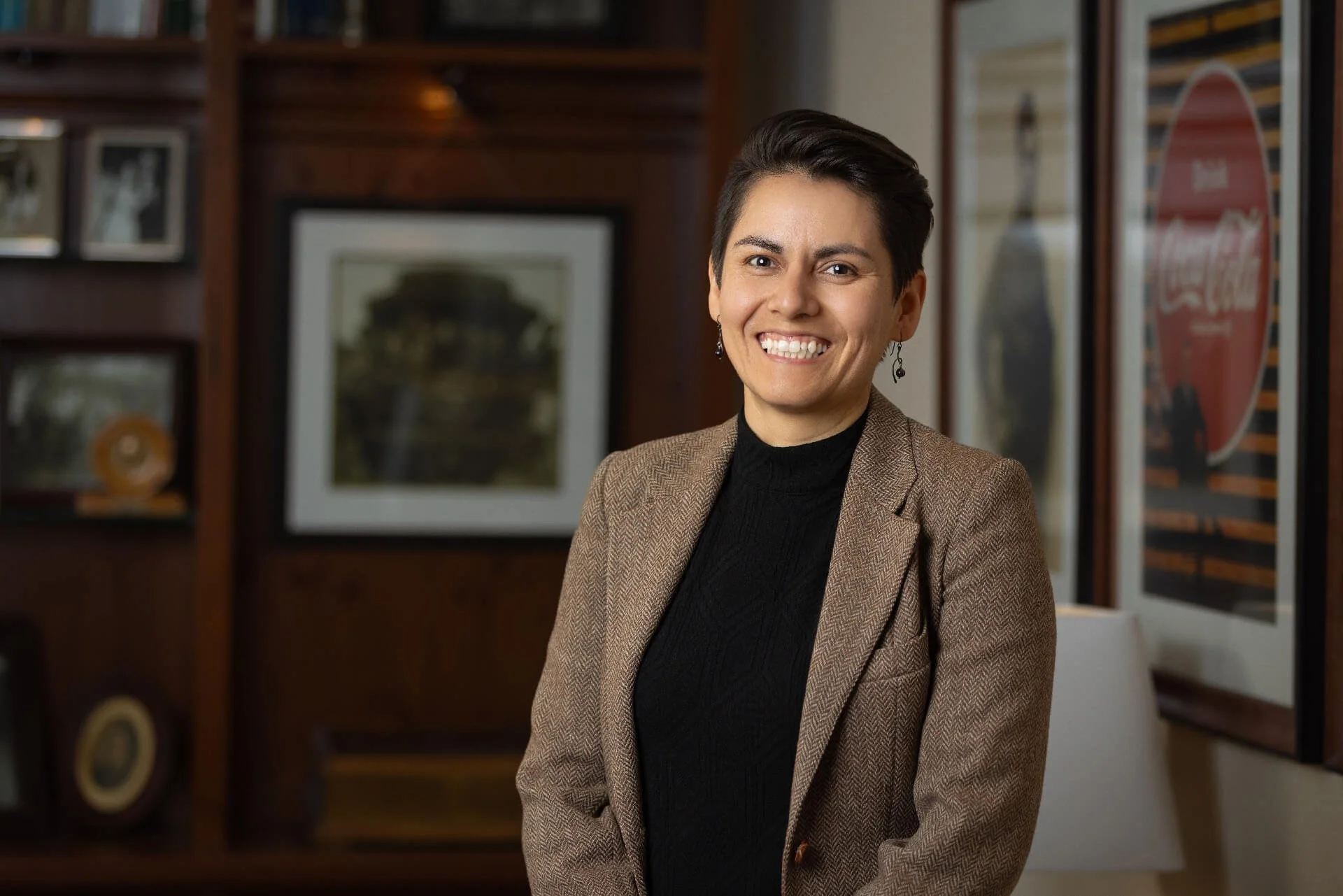 A woman smiling and wearing a brown blazer and black top inside a room with wooden paneling and framed pictures on the wall.