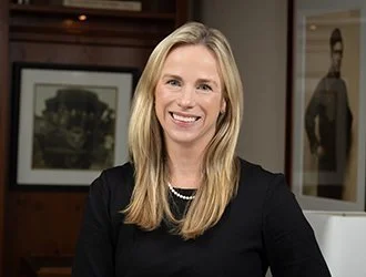 Portrait of a smiling woman with long blonde hair, wearing a black top and pearl necklace, in an office setting.