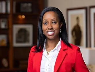A woman in a red blazer smiling in a room with framed pictures and wooden shelves.