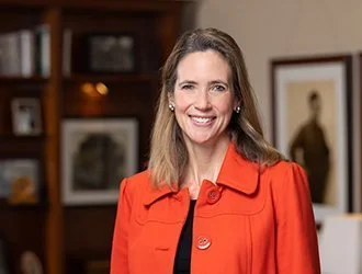 A smiling woman with light brown hair in a red blazer in an indoor setting with pictures on the wall behind her.