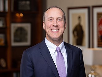 A smiling man in a dark blue suit with a white shirt and purple tie, standing indoors in front of framed artwork.