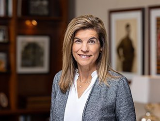 A smiling woman with long hair wearing a gray blazer and white blouse, standing in a room with framed artwork.
