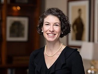 A woman with curly dark hair smiling, wearing a black top and a pearl necklace, standing indoors with framed pictures on the wall behind her.