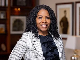 A smiling woman with curly black hair, wearing a black blouse and a white checkered blazer, standing in a room with framed artwork on the wall.