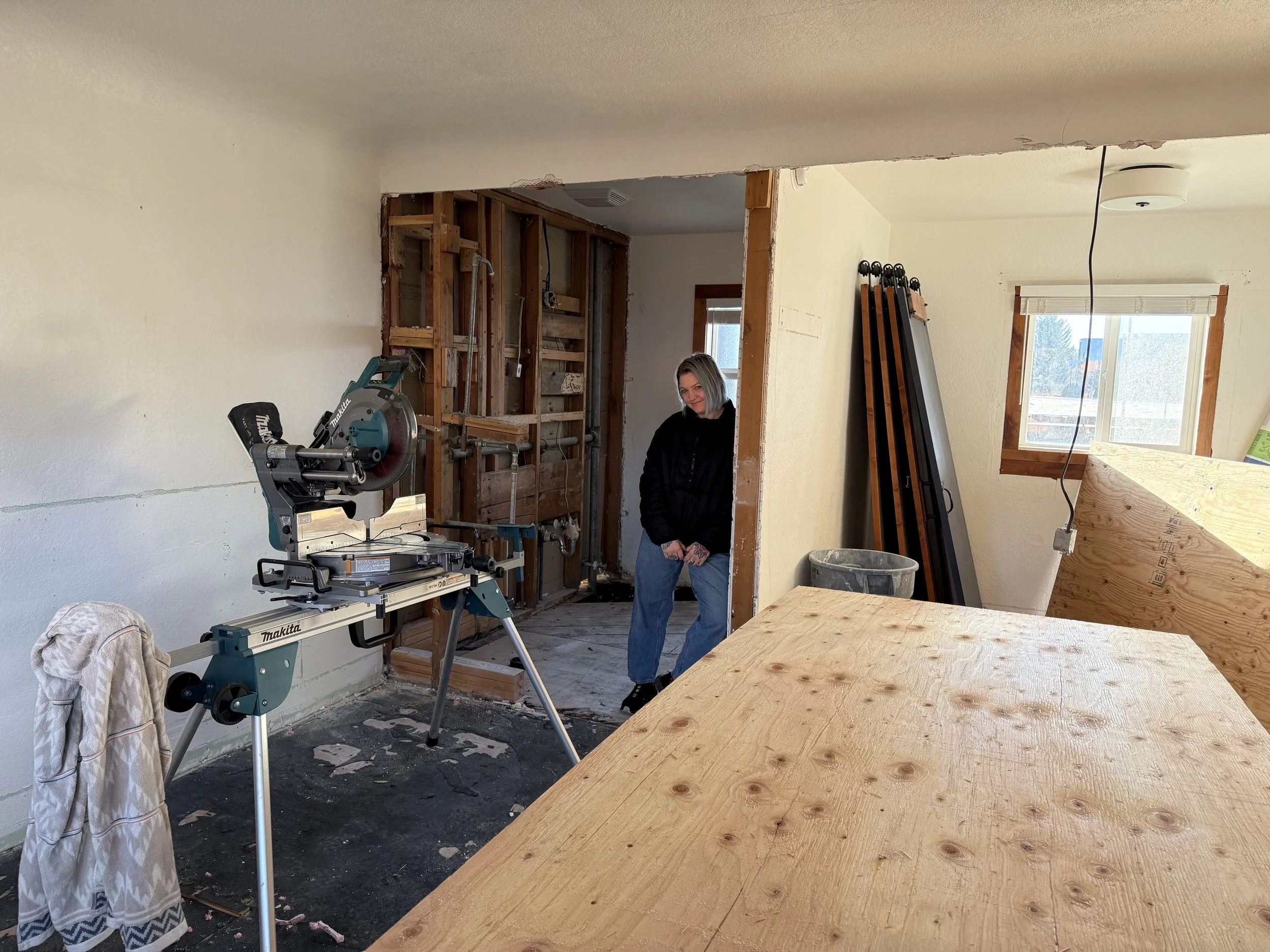 A woman standing in a room under renovation, with exposed wall framing and construction tools around, including a miter saw on a stand and wooden planks leaning against the wall.