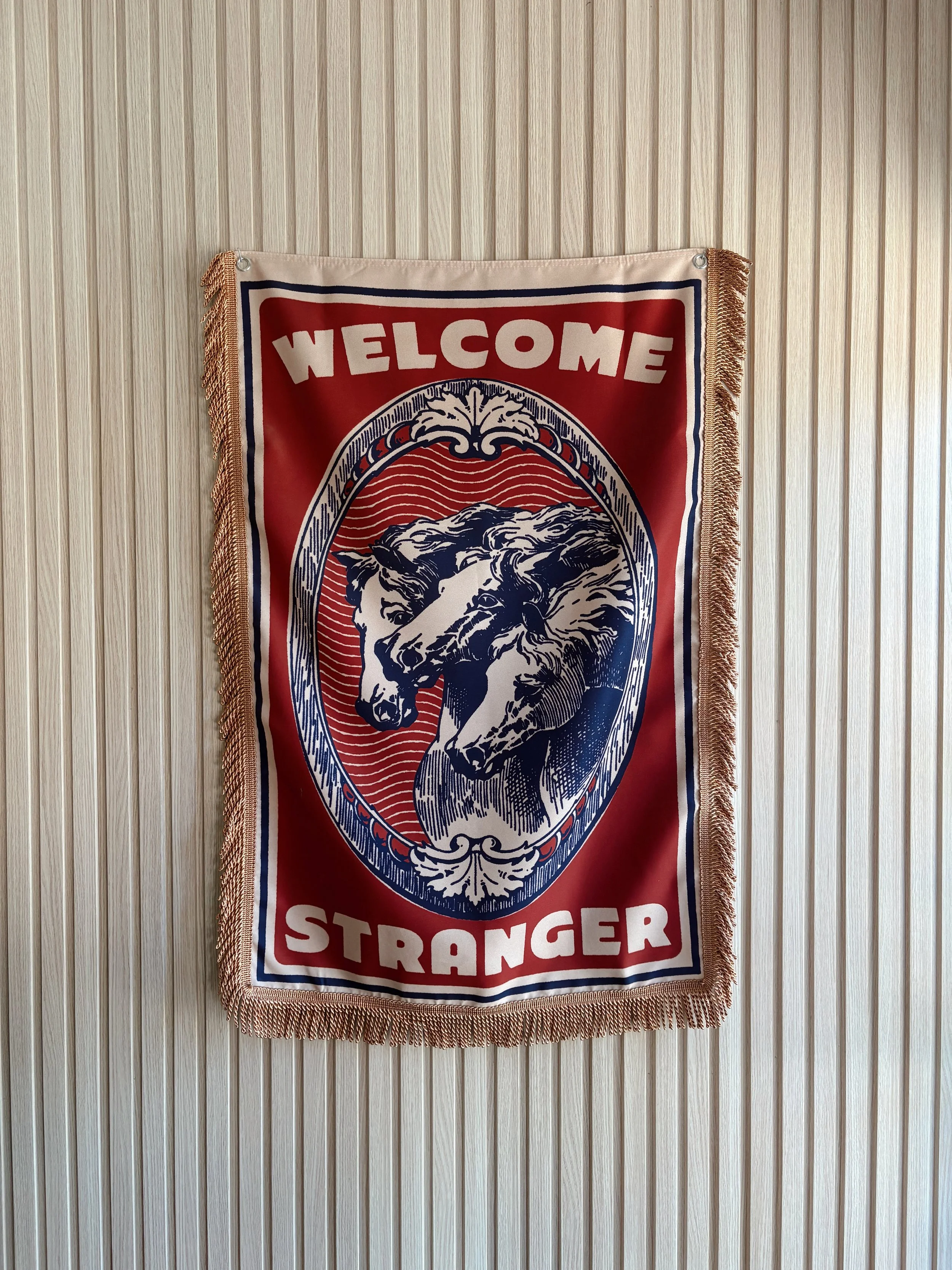A decorative wall hanging with two wolf heads facing each other, surrounded by an ornate border, with the words 'WELCOME STRANGER' at the top and bottom, hanging on a wood-paneled wall.