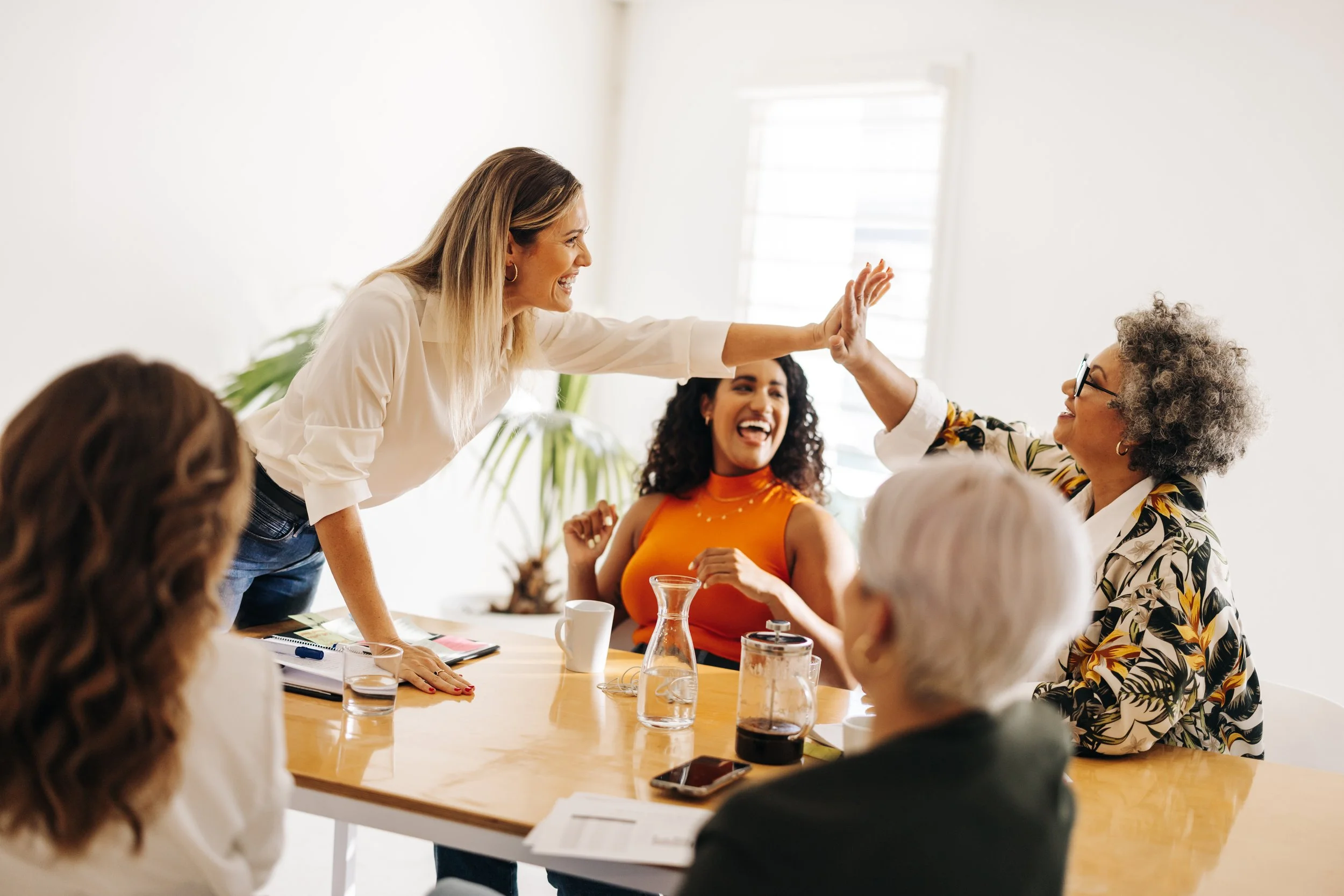 Group of women in a meeting room celebrating and giving high-fives, smiling and laughing.