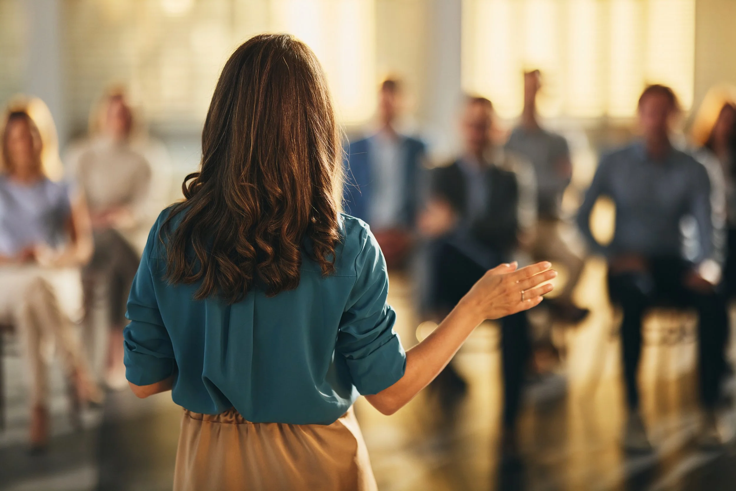A woman with brown hair wearing a blue shirt and beige pants is speaking or giving a presentation to a blurred audience in a bright room with large windows.