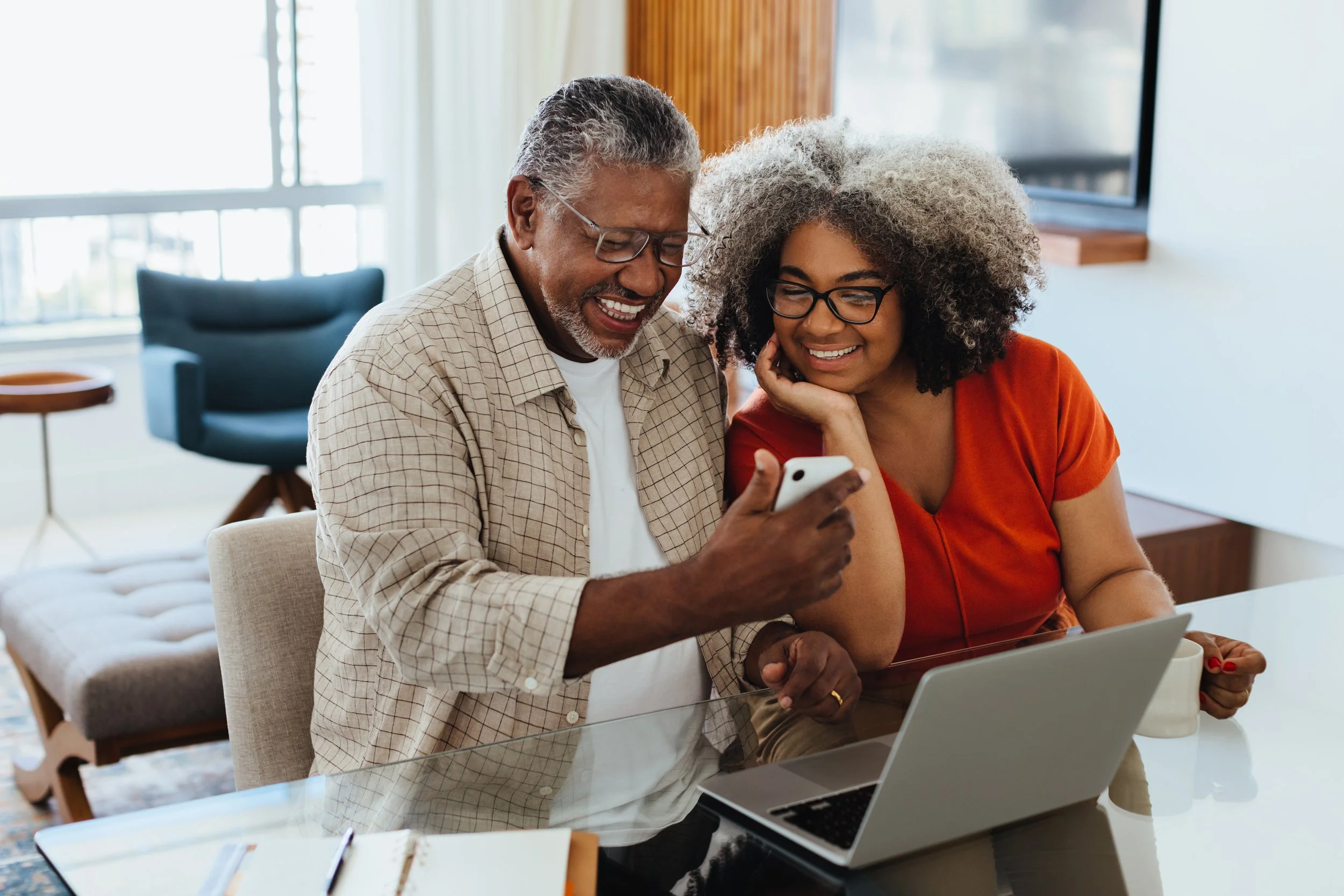 An older man and a woman sitting at a table, smiling and looking at a smartphone. There is a laptop, notebook, and cup on the table, with a window and a television in the background.