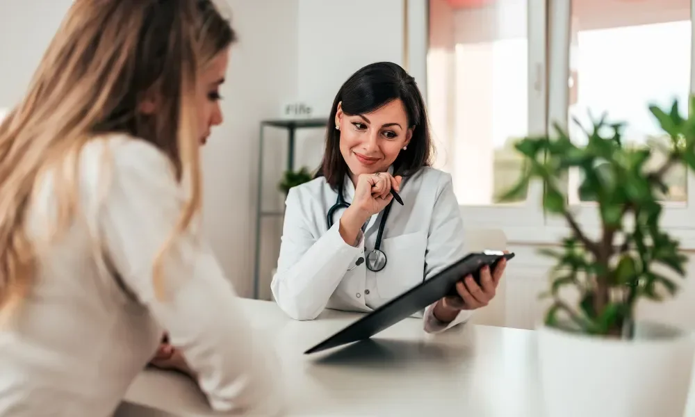 Two female doctors having a consultation in a medical office, with one holding a tablet device and the other listening attentively.