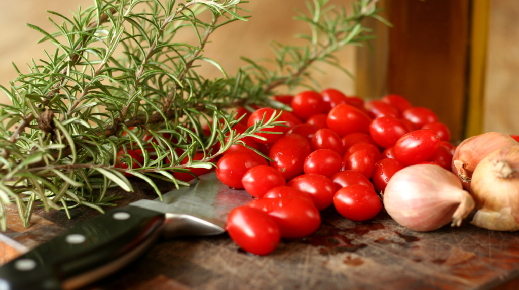 Cherry tomatoes, garlic cloves, and a sprig of rosemary on a wooden surface with a kitchen background.