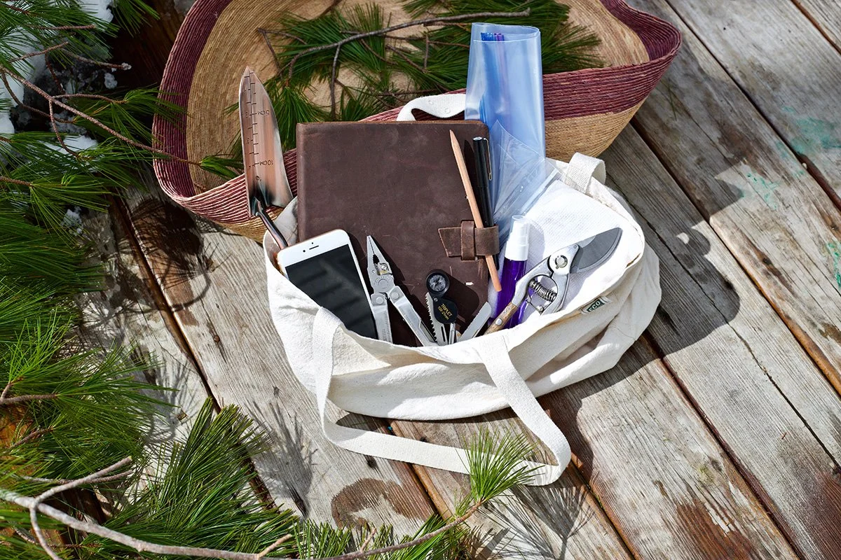 A white tote bag filled with various gardening tools and supplies on a wooden deck next to a basket with green pine branches.