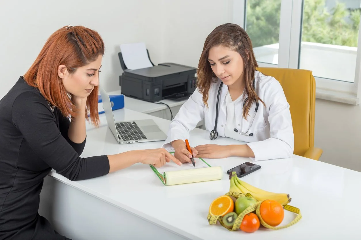 A doctor and a woman discussing healthy eating options at a table with fruits, a laptop, and a phone in an office setting.
