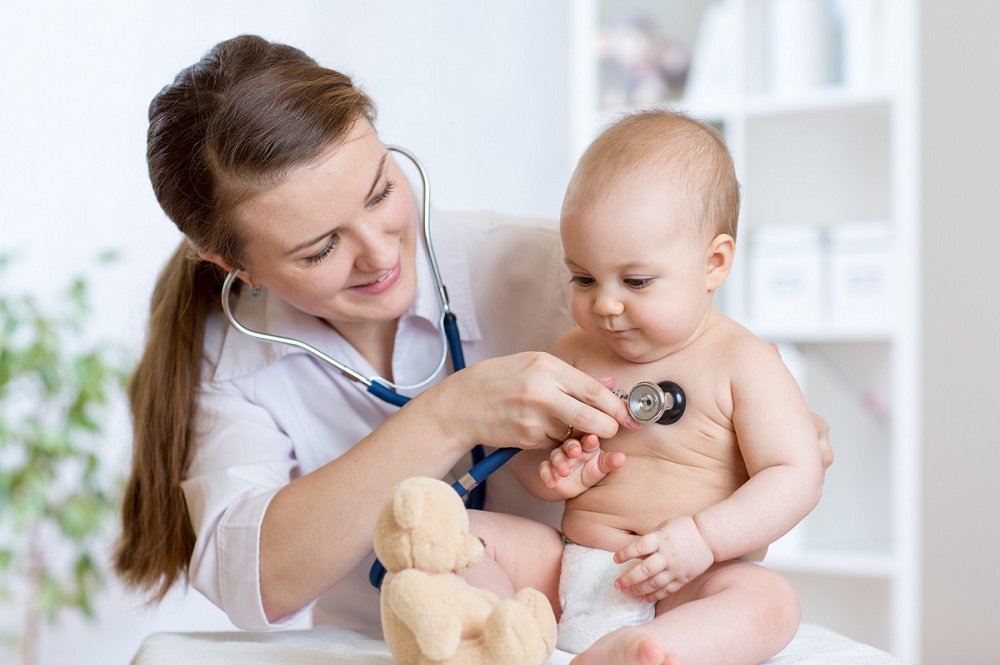 Pediatrician listening to a baby boy's chest with a stethoscope in a bright medical office.