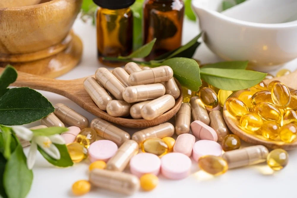 Assorted herbal supplements, capsules, and pills on a white surface with green leaves, flowers, and small medicine bottles in the background.