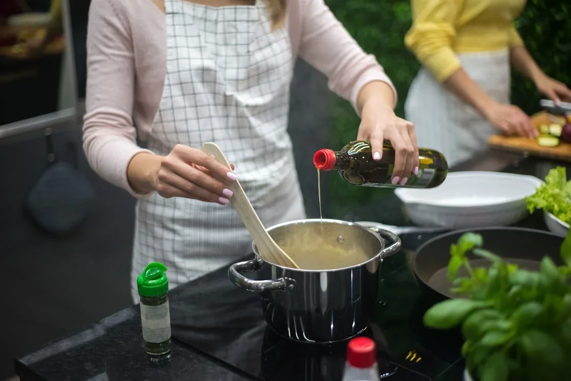 A woman in an apron cooking over a stove, pouring oil into a pot, with vegetables and cooking utensils nearby.