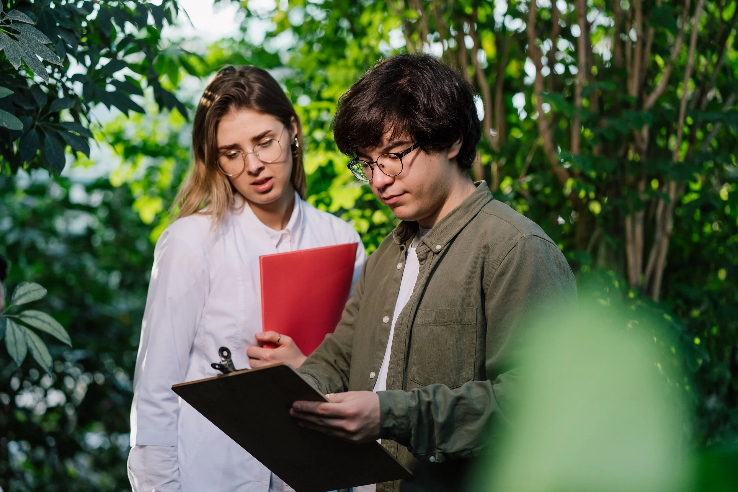 Two people, a man and a woman, standing outdoors among green trees, looking at a clipboard the man is holding.