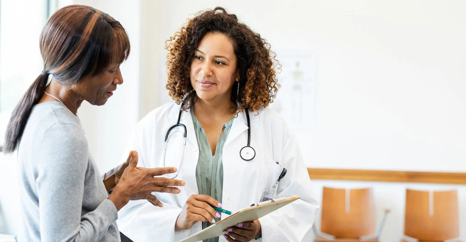 Doctor talking to patient in medical office, woman with dark hair, wearing white coat, holding clipboard, indoor clinic setting.