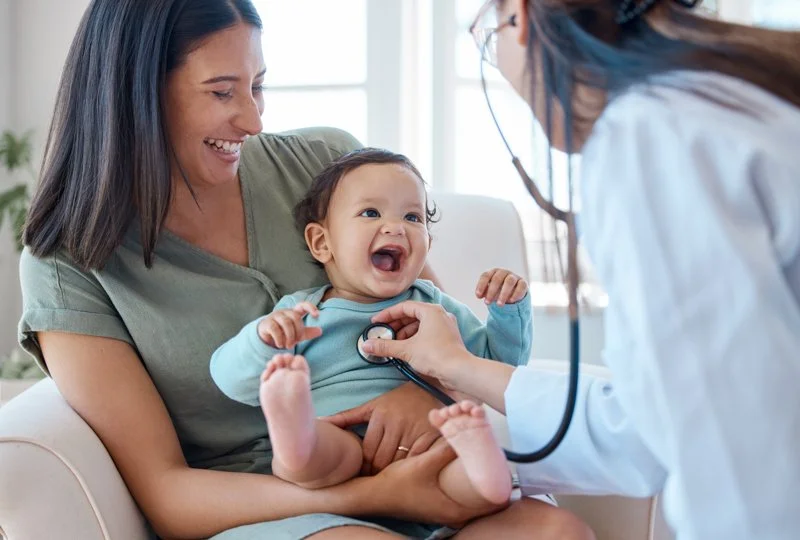 A smiling woman holding a laughing toddler during a pediatric checkup with a doctor examining the child's chest with a stethoscope.