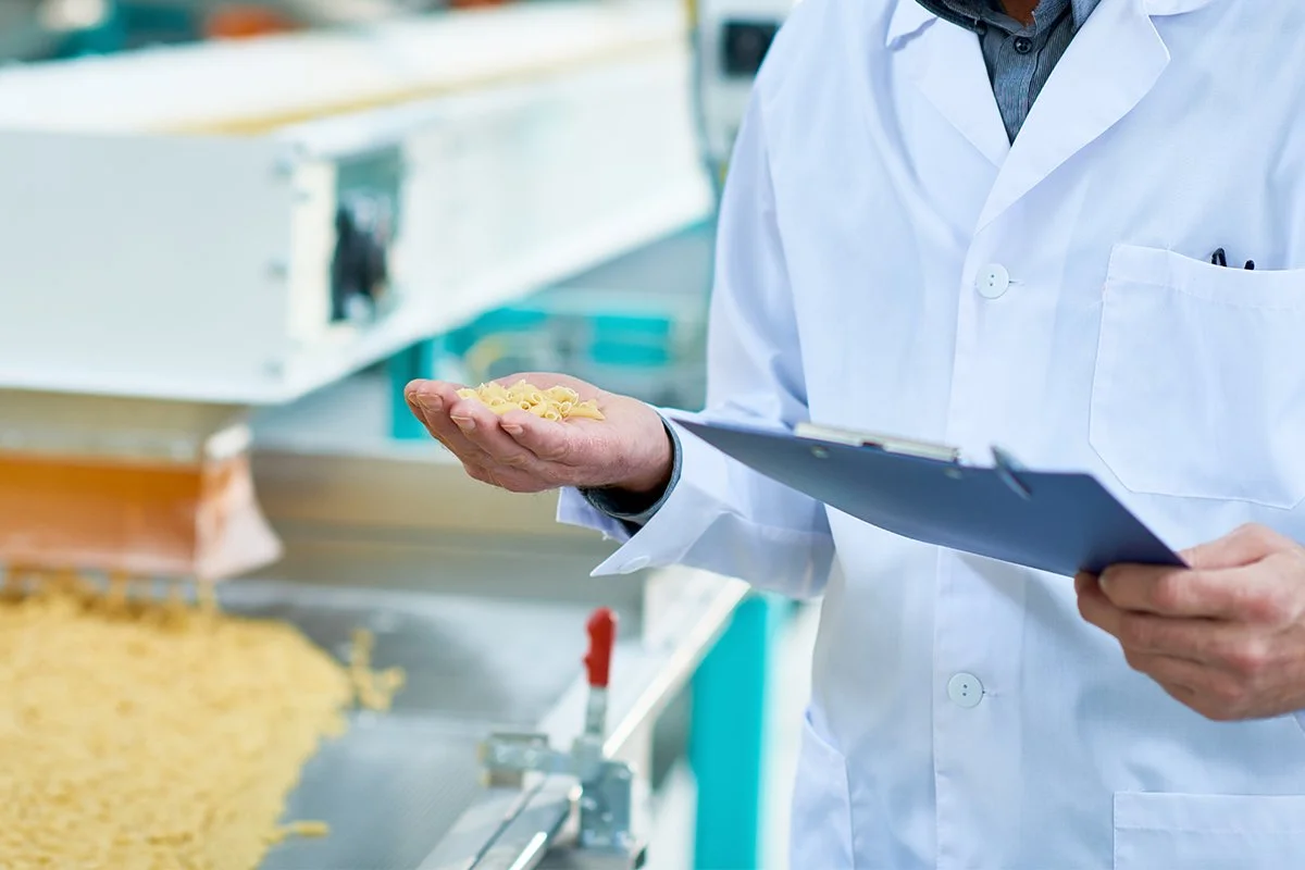 A scientist in a white lab coat holding a clipboard and examining a sample of pasta in a manufacturing or laboratory setting.