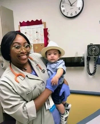 A healthcare worker holding a young child in a medical office. The woman is smiling and wearing glasses and a stethoscope. The child is wearing a hat and blue clothes. There is a clock and a blood pressure monitor on the wall behind them.