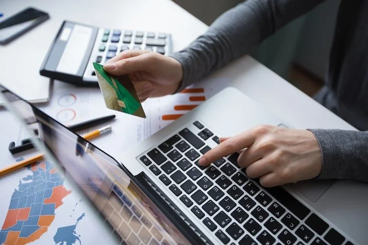 Person using a laptop and holding a credit card over financial documents and calculator on a desk.