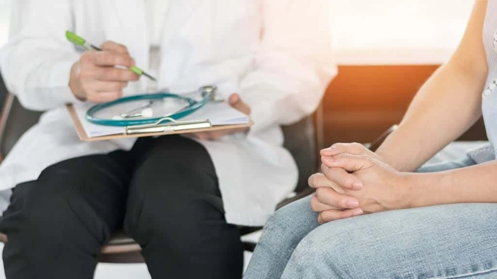 A doctor holding a clipboard and pen, sitting on a chair, talking to a patient whose hands are clasped together in her lap.