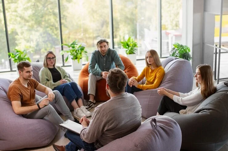 A group therapy session with six adults seated on colorful couches in a circle in a bright room with large windows and green plants.