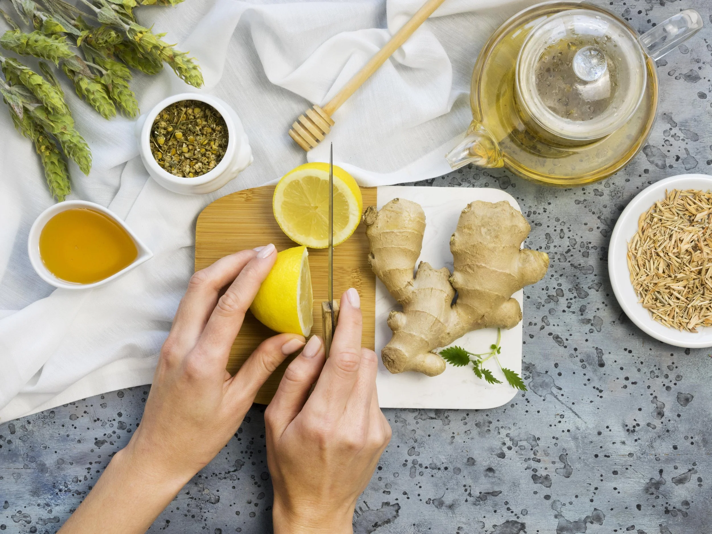 Person slicing lemon on a cutting board surrounded by fresh ginger, honey, herbs, and tea on a gray speckled surface.