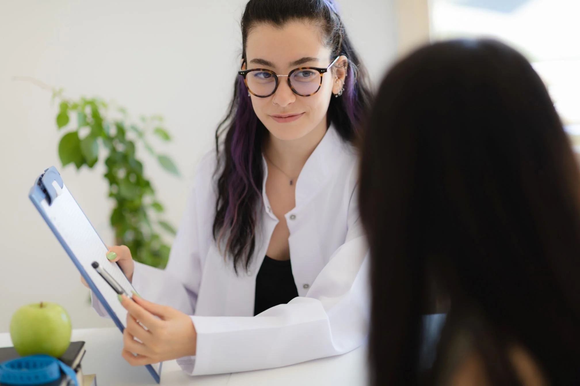 A woman with glasses and dark hair with purple streaks, wearing a white lab coat, sitting at a desk with a clipboard, talking to another person whose back is to the camera.