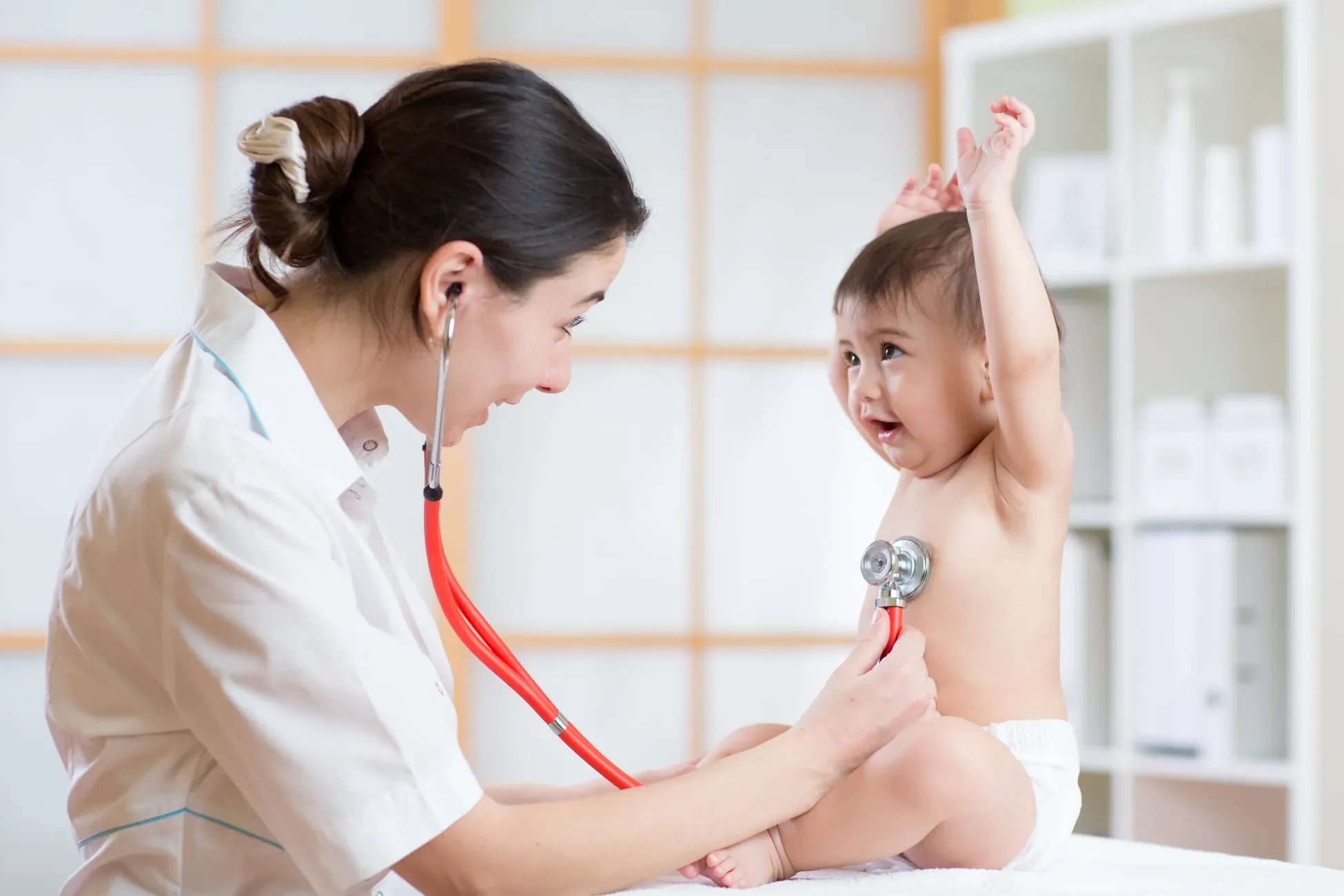 A female doctor using a stethoscope to examine a happy, shirtless toddler sitting on an examination table, raising one arm.