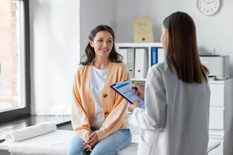 A woman in her 20s sitting on an examination table in a doctor’s office, smiling and talking to a female doctor holding a clipboard.