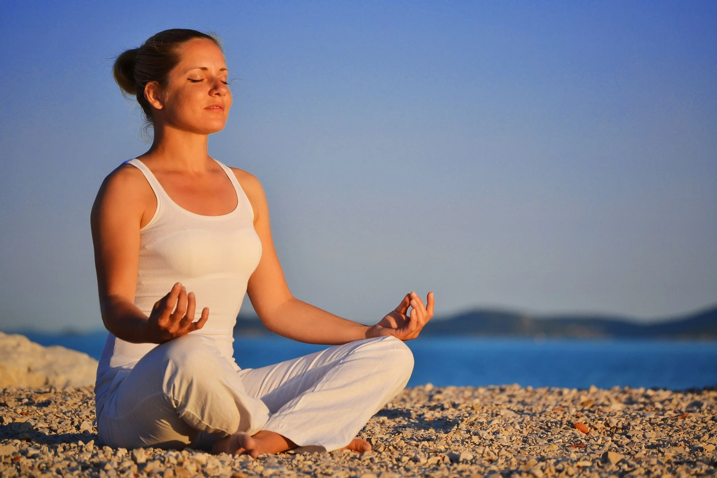 A woman sitting cross-legged on a sandy beach meditating with her eyes closed, holding her hands in a mudra, in the late afternoon or early evening sunlight with a calm sea and distant islands in the background.