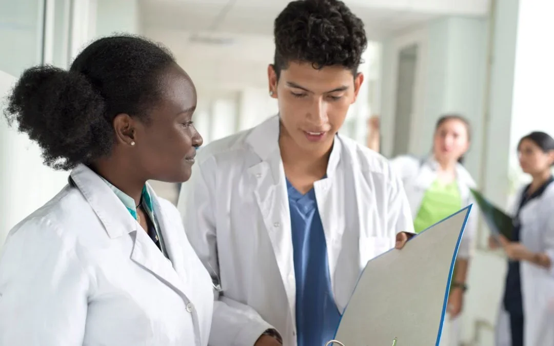 Two young medical professionals in white coats discussing a folder in a hospital corridor, with three other people in medical attire in the background.
