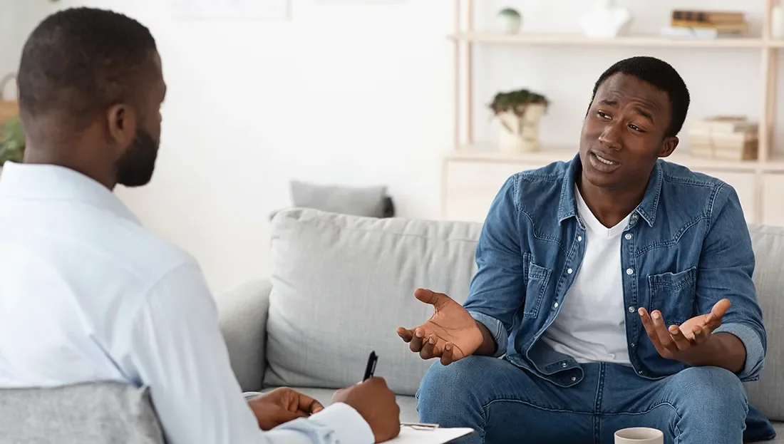 A young man sitting on a sofa, talking and gesturing with his hands to a counselor or therapist who is taking notes.