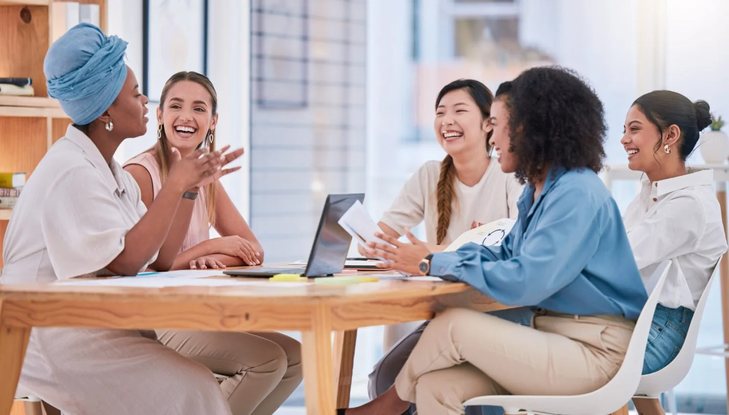 Five women sitting around a wooden table, engaged in a lively discussion and sharing laughs, in a bright room with large windows.